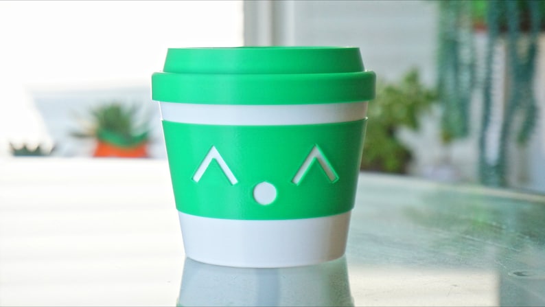 May include: A green and white reusable coffee cup with a cute face. The cup has a white band around the middle and a green lid.