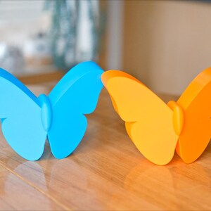 May include: Two butterfly-shaped decorative objects. One is light blue, the other is orange. They are placed on a wooden surface. The butterflies have a smooth, solid appearance, with rounded wings and a central body.