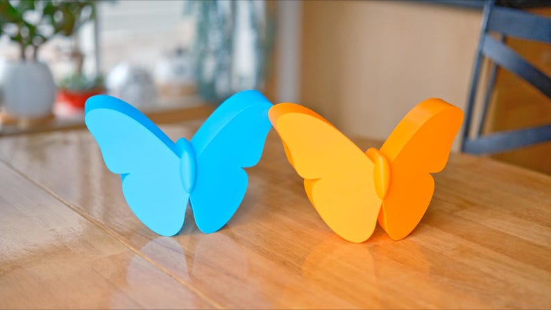 May include: Two butterfly-shaped decorative objects. One is light blue, the other is orange. They are placed on a wooden surface. The butterflies have a smooth, solid appearance, with rounded wings and a central body.