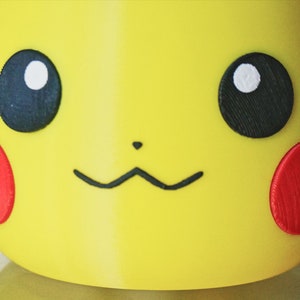 May include: Close-up of a yellow Pikachu character with black eyes, a black nose, and red cheeks. The character is made of plastic and has a 3D appearance.