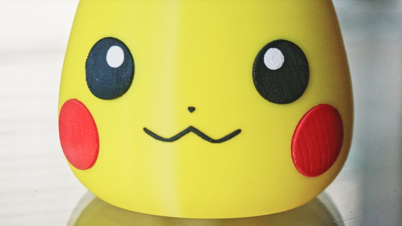 May include: Close-up of a yellow Pikachu character with black eyes, a black nose, and red cheeks. The character is made of plastic and has a 3D appearance.
