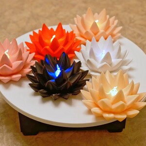May include: Six lotus-shaped candles with different colored lights, arranged in a circle on a white platform. The candles are pink, orange, white, black, and two shades of peach.