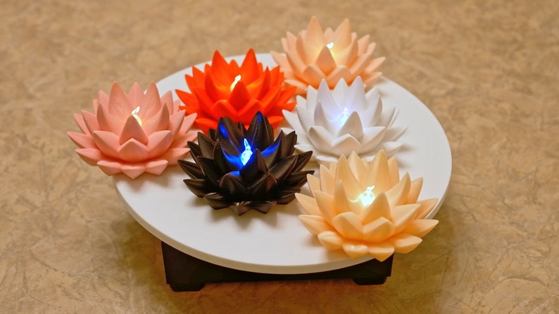 May include: Six lotus-shaped candles with different colored lights, arranged in a circle on a white platform. The candles are pink, orange, white, black, and two shades of peach.
