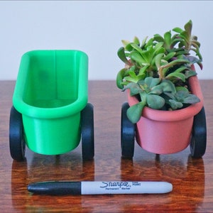 May include: Two small green and terracotta planters on wheels, each with a different type of succulent plant. The planters are on a wooden surface with a black marker pen.