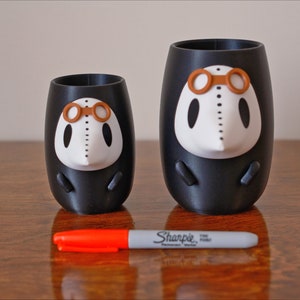 May include: Two black and white 3D printed cups with a white face and brown goggles. The cups are different sizes and are sitting on a wooden surface.