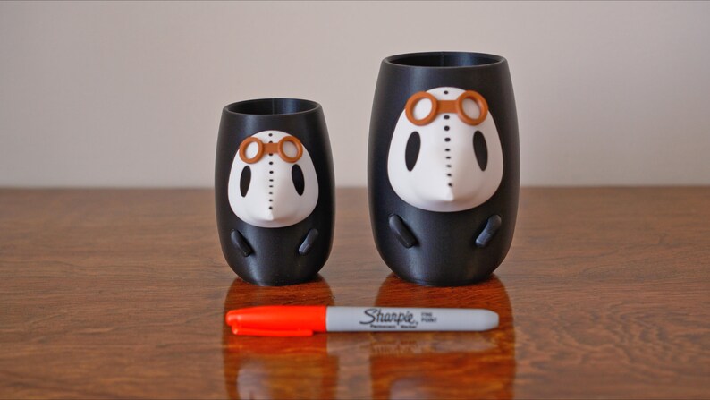 May include: Two black and white 3D printed cups with a white face and brown goggles. The cups are different sizes and are sitting on a wooden surface.