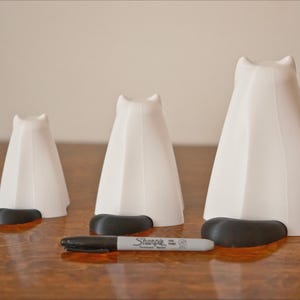 May include: Three white ceramic cat figurines of varying sizes, each with a black base. The figurines are arranged in a row on a wooden surface.