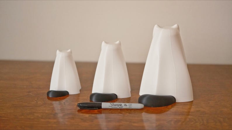 May include: Three white ceramic cat figurines of varying sizes, each with a black base. The figurines are arranged in a row on a wooden surface.