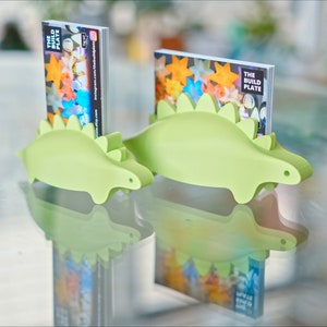 May include: Two green dinosaur-shaped business card holders, one larger than the other, are on a glass table. The larger holder has a business card with the text "The Build Plate" in it. The smaller holder has a business card with the text "The Build Plate" in it.