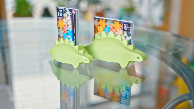 May include: Two green dinosaur-shaped business card holders, one larger than the other, are on a glass table. The larger holder has a business card with the text "The Build Plate" in it. The smaller holder has a business card with the text "The Build Plate" in it.