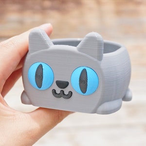 Pode incluir: Um vaso de planta cinza em forma de gato com olhos azuis e uma boca sorridente. O vaso &eacute; feito de pl&aacute;stico impresso em 3D.