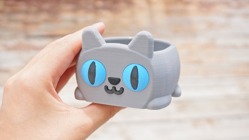 Pode incluir: Um vaso de planta cinza em forma de gato com olhos azuis e uma boca sorridente. O vaso &eacute; feito de pl&aacute;stico impresso em 3D.