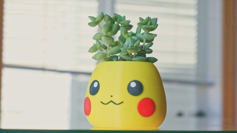 May include: A yellow ceramic planter shaped like a cartoon character with a green succulent plant growing inside. The planter has a smiling face with black eyes and a red nose.