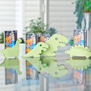 May include: Four green dinosaur-shaped card holders on a glass table. Each holder has a card with a colourful design and the text "The Big Plate".