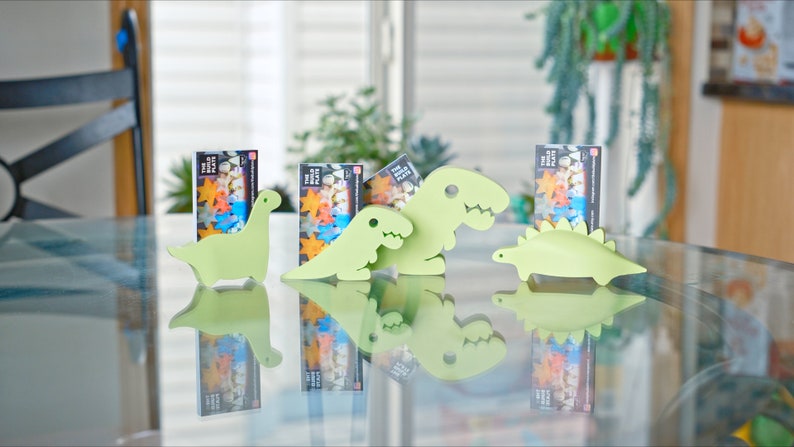 May include: Four green dinosaur-shaped card holders on a glass table. Each holder has a card with a colourful design and the text "The Big Plate".