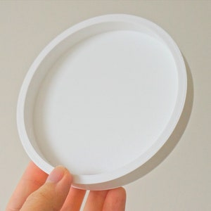 May include: A white round ceramic tray with a raised rim. The tray is empty and appears to be made of a smooth material.