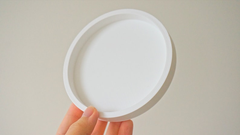 May include: A white round ceramic tray with a raised rim. The tray is empty and appears to be made of a smooth material.