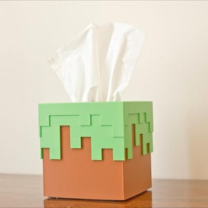 A cube tissue box cover inspired by the dirt grass block from the popular video game, Minecraft, is on a wooden table with a white background. It has bright green pixelated grass at the top half and the bottom half is brown.