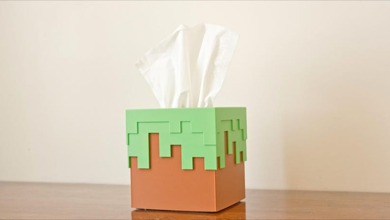 A cube tissue box cover inspired by the dirt grass block from the popular video game, Minecraft, is on a wooden table with a white background. It has bright green pixelated grass at the top half and the bottom half is brown.