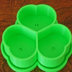 May include: A bright green, three-leaf clover-shaped cookie cutter. The cutter is made of plastic and has a smooth surface. Each leaf has a small hole in the center. The cutter is designed for making cookies or other treats in the shape of a clover.