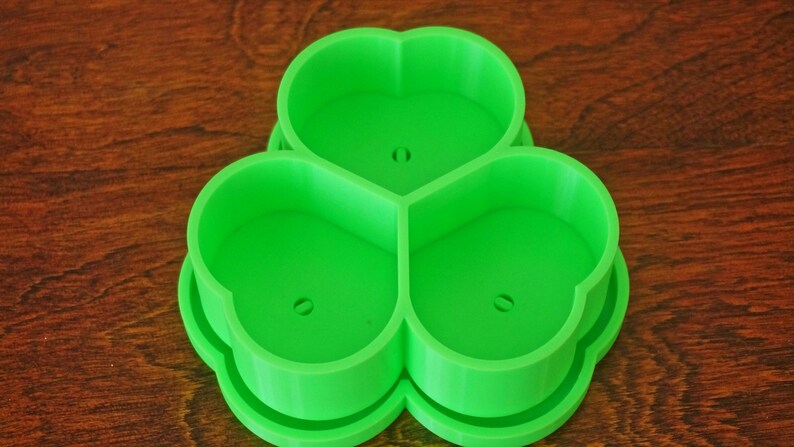 May include: A bright green, three-leaf clover-shaped cookie cutter. The cutter is made of plastic and has a smooth surface. Each leaf has a small hole in the center. The cutter is designed for making cookies or other treats in the shape of a clover.