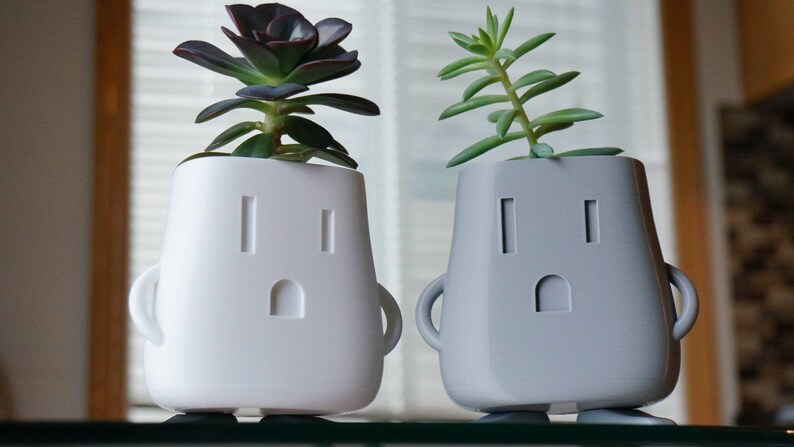 Op de afbeelding: Twee witte en grijze keramische potten in de vorm van een stopcontact met een gezicht. De potten hebben kleine armen en benen. De witte pot heeft een succulent met donkergroene bladeren. De grijze pot heeft een succulent met lichtgroene bladeren.