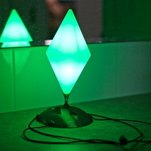 May include: A green, diamond-shaped lamp with a black base and a long cord. The lamp is illuminated, emitting a bright green glow. A smaller, triangular lamp is reflected in the background. The overall theme is modern lighting.