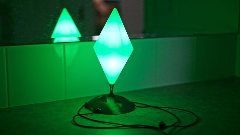 May include: A green, diamond-shaped lamp with a black base and a long cord. The lamp is illuminated, emitting a bright green glow. A smaller, triangular lamp is reflected in the background. The overall theme is modern lighting.