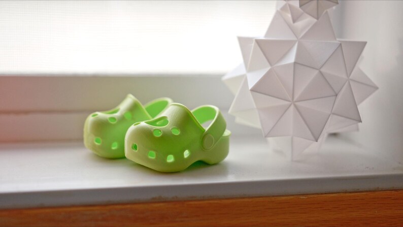 May include: Two bright green plastic clogs, one slightly larger than the other, are sitting on a white surface.