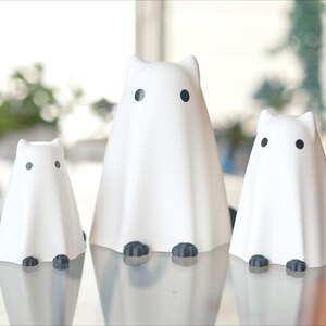 May include: Three white ceramic ghost figurines with black eyes and feet. The ghosts are arranged in a row on a glass table.
