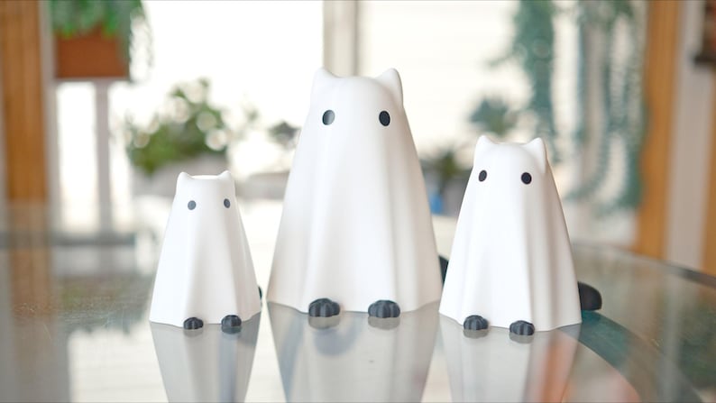 May include: Three white ceramic ghost figurines with black eyes and feet. The ghosts are arranged in a row on a glass table.