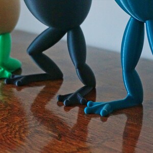 May include: Three frog-shaped figurines with different colored legs. The first frog has green legs, the second has black legs, and the third has blue legs.
