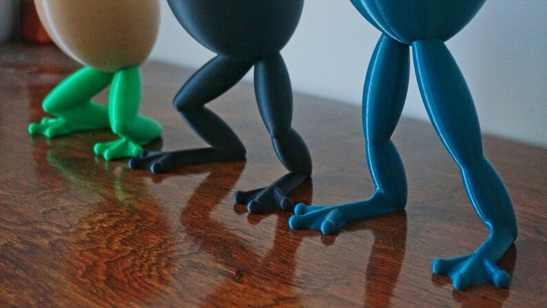 May include: Three frog-shaped figurines with different colored legs. The first frog has green legs, the second has black legs, and the third has blue legs.