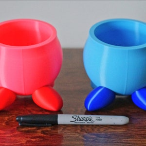 May include: Two small, 3D printed planters with legs. One planter is pink with red legs, and the other is blue with blue legs.