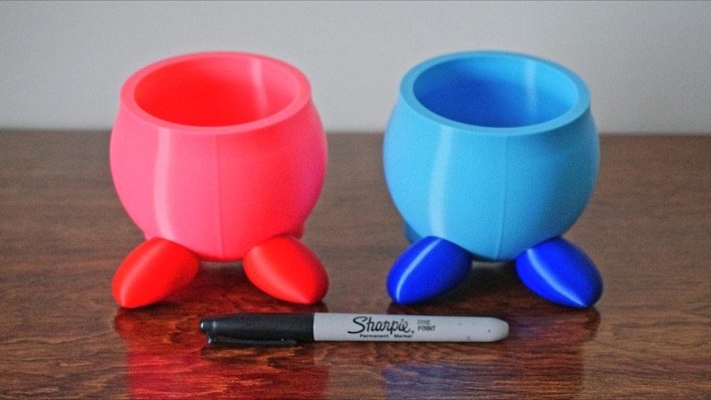 May include: Two small, 3D printed planters with legs. One planter is pink with red legs, and the other is blue with blue legs.