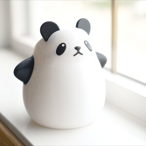 Op de afbeelding: Een wit keramiek panda-beeldje met zwarte oren, ogen, neus en mond. De panda staat op een wit oppervlak.
