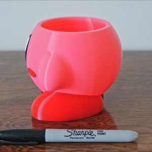 Puede incluir: Maceta de impresi&oacute;n 3D rosa y roja con forma del personaje Kirby de la serie de videojuegos Kirby. La maceta est&aacute; sobre una superficie de madera.