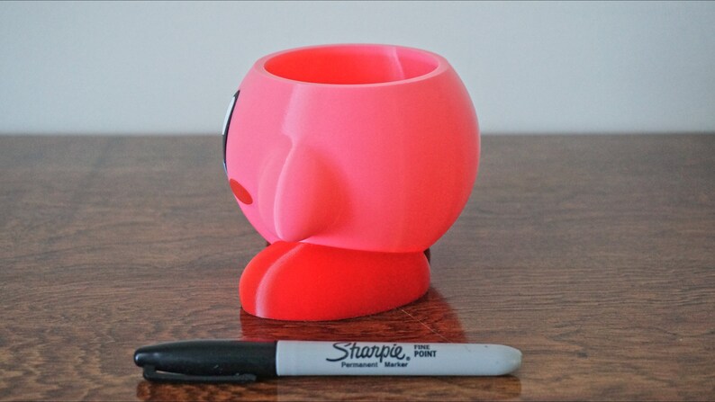 Puede incluir: Maceta de impresi&oacute;n 3D rosa y roja con forma del personaje Kirby de la serie de videojuegos Kirby. La maceta est&aacute; sobre una superficie de madera.