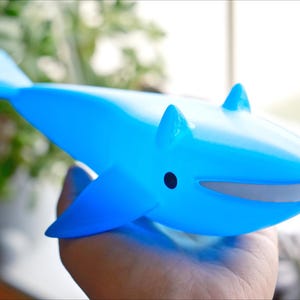 May include: A light blue whale-shaped toy with fins and a small black eye. The toy has a gray mouth and is held in a hand. The background is blurred, with green foliage visible.