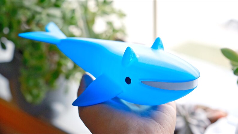 May include: A light blue whale-shaped toy with fins and a small black eye. The toy has a gray mouth and is held in a hand. The background is blurred, with green foliage visible.