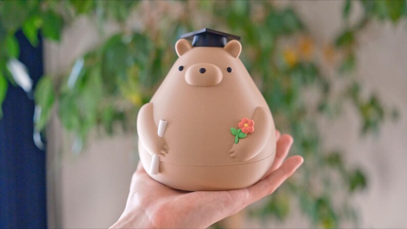 Puede incluir: Figura en forma de oso marr&oacute;n claro, con birrete negro, diploma y flor. La figura es redonda y lisa, con peque&ntilde;os ojos negros y nariz de bot&oacute;n. El oso est&aacute; en la mano de una persona.