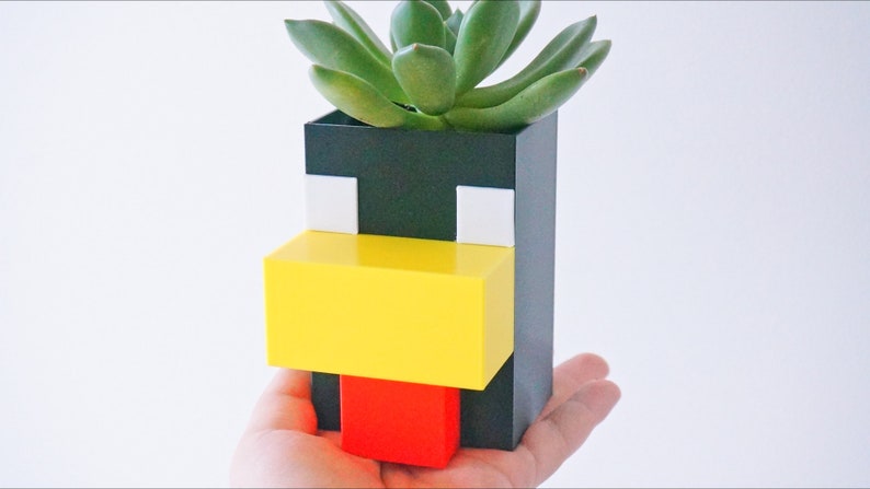 May include: A black, white, yellow, and red planter shaped like a pixelated face with a succulent plant growing inside.