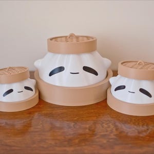 Puede incluir: Tres figuritas de cer&aacute;mica blanca con forma de alb&oacute;ndigas con tapas marrones y ojos negros. Las figuritas est&aacute;n dispuestas en fila sobre una superficie de madera.