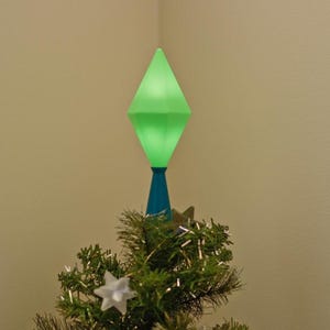 May include: A green, diamond-shaped Christmas tree topper with a blue base, illuminated with a soft green glow. The topper sits atop a small, decorated Christmas tree with white star ornaments and silver tinsel.