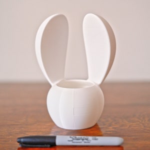 May include: White ceramic bunny-shaped planter with two upright ears. The planter has a round base and a cylindrical opening for planting.