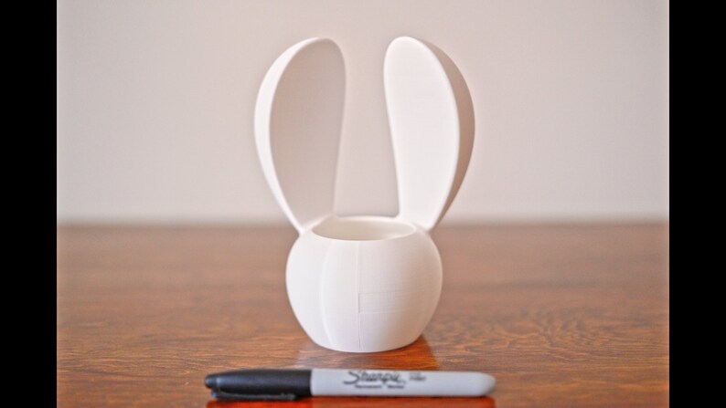 May include: White ceramic bunny-shaped planter with two upright ears. The planter has a round base and a cylindrical opening for planting.