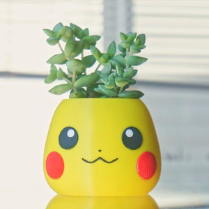 May include: A yellow ceramic planter shaped like a cartoon character with red cheeks and black eyes. A small green succulent plant is growing out of the top of the planter.