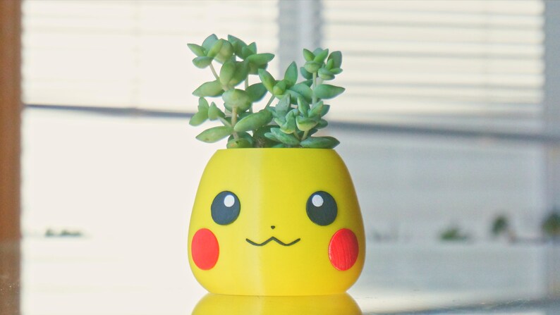 May include: A yellow ceramic planter shaped like a cartoon character with red cheeks and black eyes. A small green succulent plant is growing out of the top of the planter.