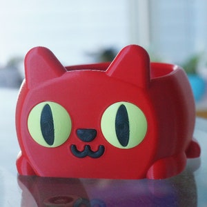 Pode incluir: Um vaso vermelho em forma de gato com olhos verdes e um nariz e boca pretos. O vaso &eacute; feito de pl&aacute;stico e tem uma superf&iacute;cie lisa.