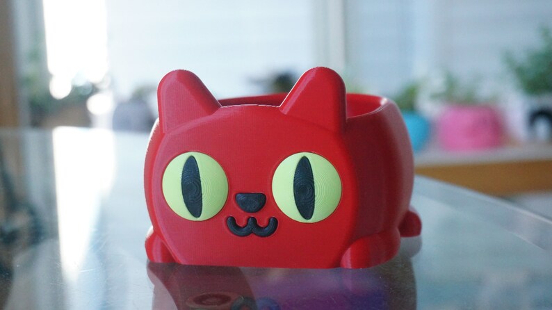 Pode incluir: Um vaso vermelho em forma de gato com olhos verdes e um nariz e boca pretos. O vaso &eacute; feito de pl&aacute;stico e tem uma superf&iacute;cie lisa.